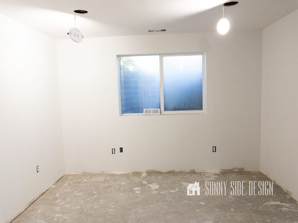 Empty basement room with white primer on walls and a cement floor, with 1 large window.