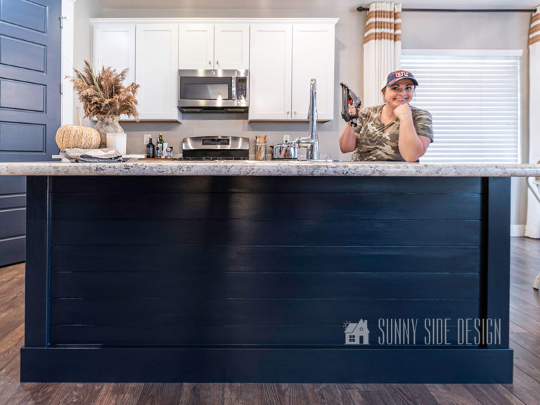 Woman holding nail gun with a camo t-shirt at kitchen island after makeover with shiplap and trim, painted navy blue. White shaker cabinets in background, with a navy blue pantry door.