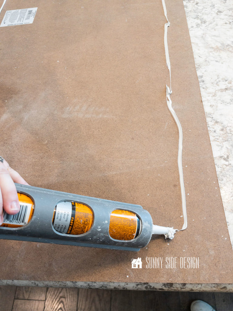 Woman applies construction adhesive to the backside of the shiplap panel to adhere it to the back of the kitchen island.
