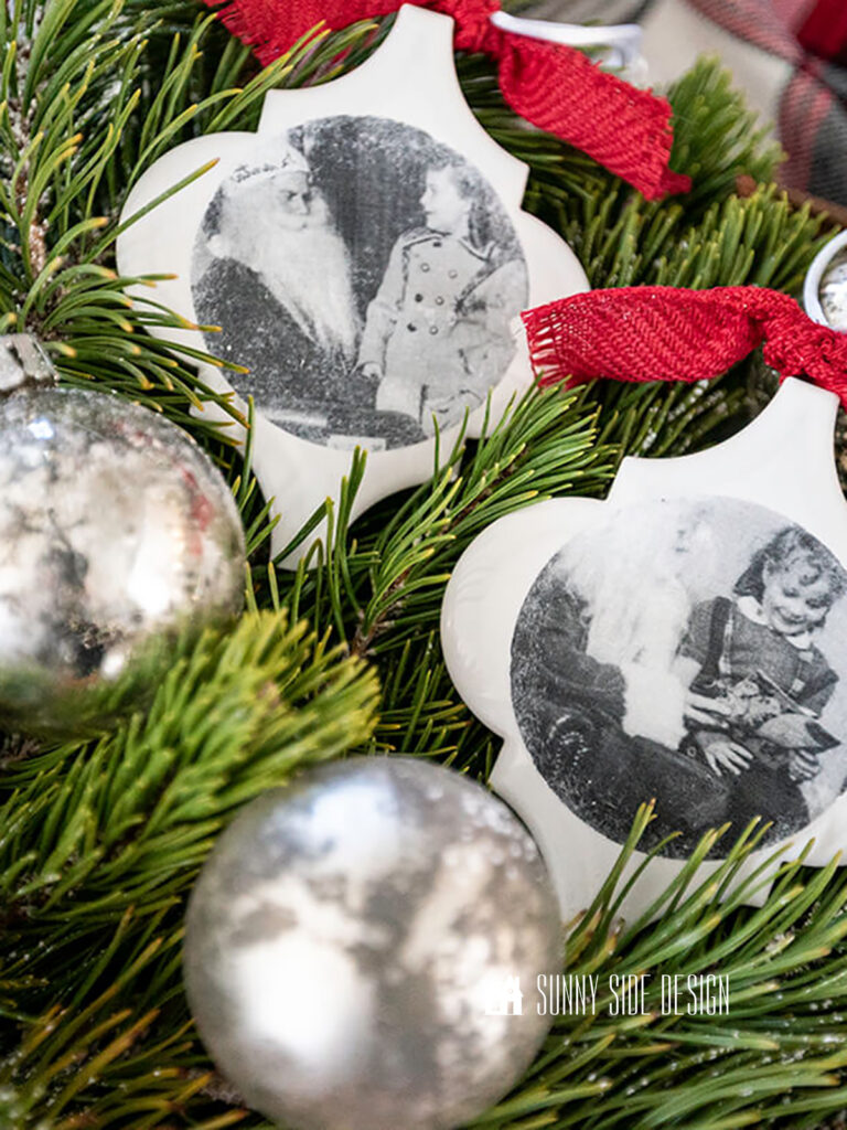 Personalized Christmas ornaments made with white arabeque tiles and old black and white photos placed in a bed of fresh greenery and mercury glass ornaments with a red plaid flannel cloth.