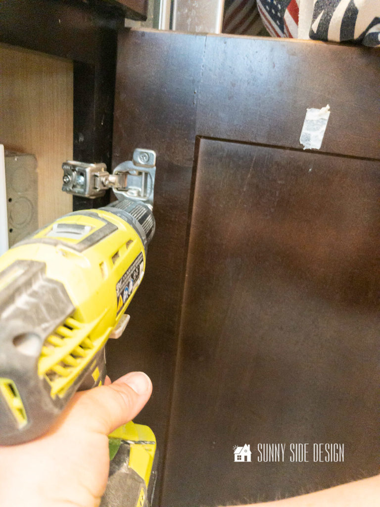 Woman's hand holding drill, removing hardware from cabinet door for makeover kitchen island.