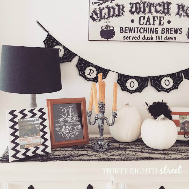 White console table decorated with black spider web table runner, white lamp with black lamp shade, framed diy printable art, candleabra, and a "spooky" bunting draped on the wall, white pumpkins. easy Halloween decor ideas for your home.