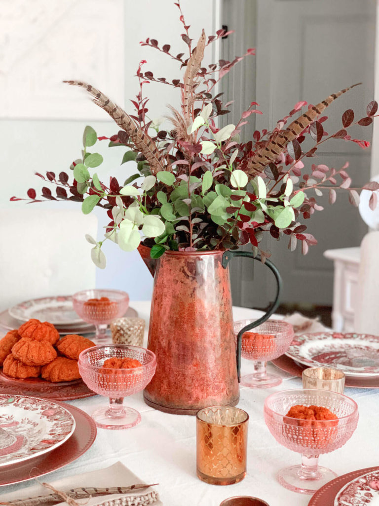 Fall table decor, Copper pitcher if fulled with burgundy, green eucaluptus and pheasant feathers. At each place setting is a burgundy charger topper with a burgundy floral plate. Rose hue gobletw filled ith a pumpkin cake are also set at each placesetting.