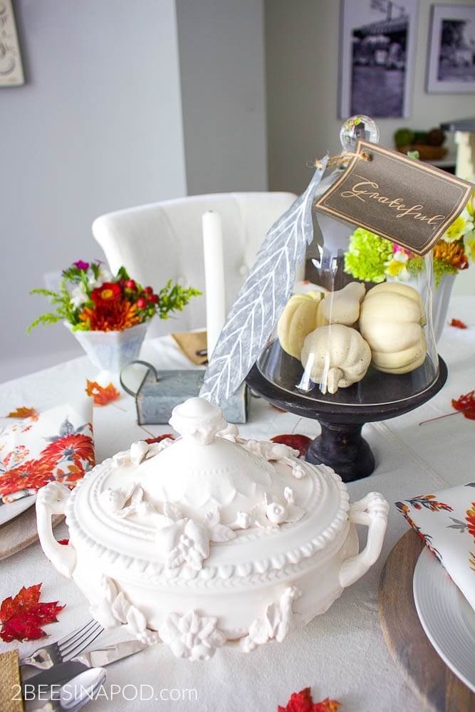 Bright and colorful Fall tablescape with red maple leaves scattered on a white linen tablecloth, white milkglass is filled with red and orange hue fresh flowers and greenery. A cloche if fulled with white pumpkins while a galvanized metal leaf it tied to the top with a card with the word grateful. White candles, and a beauitful white floral tureen.