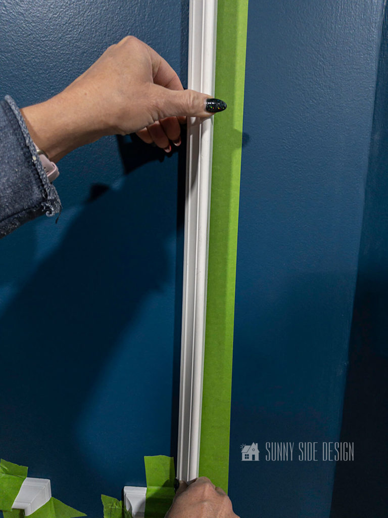 Woman places white picture frame moulding into position on blue wall aligning with green painters tape.