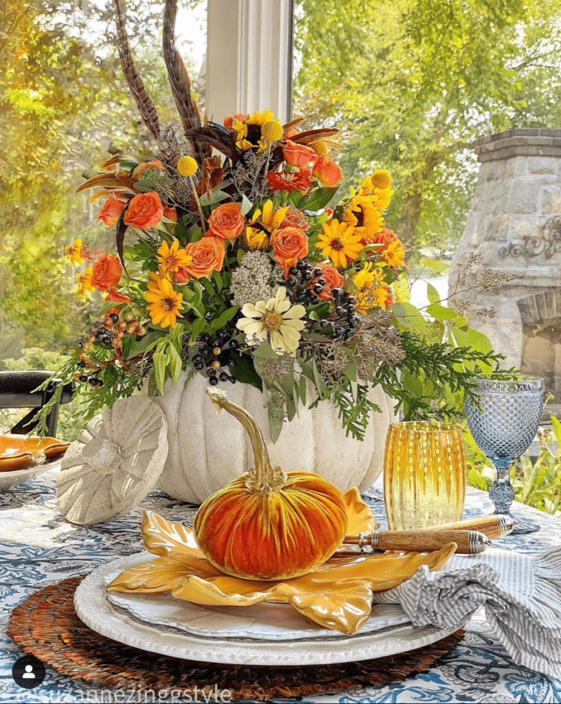 Outdoor fall tablescape idrad with a woven placemat, white charger topped with a white plate topped with a yellow leaf plate, amber and blue glassware is set at each placesetting, In the center of the table is a white pumpkin contain filled with yellow and orange hue flowers, dark purple berries and feathers. At each place setting is also a velvet orange pumpkin.