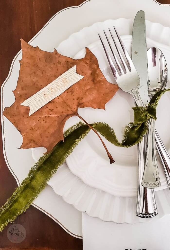 Natural leaf place card with a name stamped on a white twill ribbon placed on a maple leaf, on white dinnerware, topped with silverware tied with green fringed ribbon.