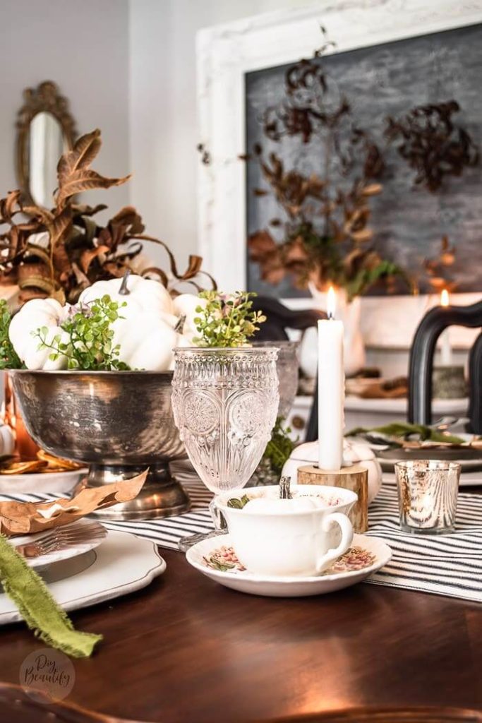 Vintage solver bowl filled with greens, white pumpkins and brown leaves setting on a black and white ticking table runner, A white mini pumpkin is placed in a floral teacup at each place setting. White chargers with white dinnerware topped with silverware tied with an avocado ribbon.