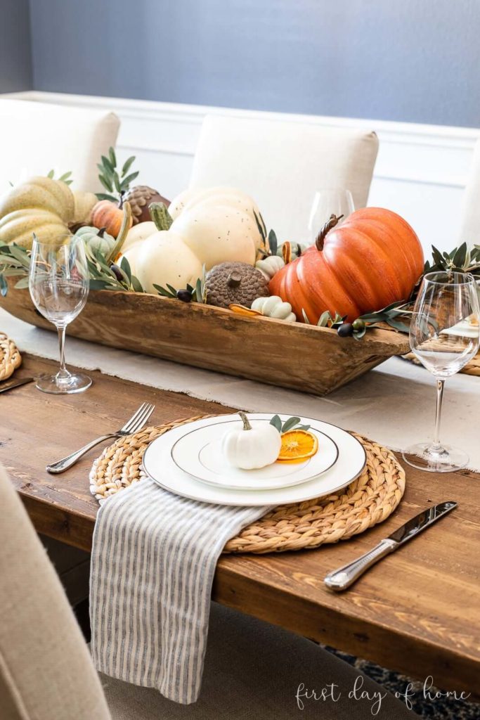 Fall tablescape with a natural linen tablerunner, woven placemats topped with a blue and white striped napkin, topped with white silver rimmed dinnerware, topped with a white pumpkin and a dried orange slice. The centerpieces is an old wooden dough bowl filled with greenery, acorns and various colored pumkins.
