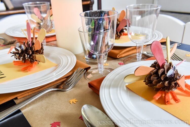 Fall tablescape idea for the kids table, Kraft paper table tunner, topped with an orange napkin, white plated, name tag and a pinecone turkey. Glass cups are filled with crayons.