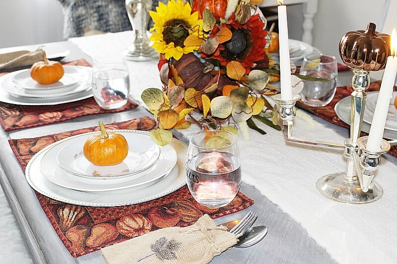 Rust colored pumpkin placemats are placed on the table for this fall tablescape, topped with white dinnerwre, topped with a mini orange pumpkin. Clear glassed and a burlap bag stamped with a leaf to hold silverware. Centerpiece of sunflowers, pumpkins and eucalyptus leaves in a woven cornicopia