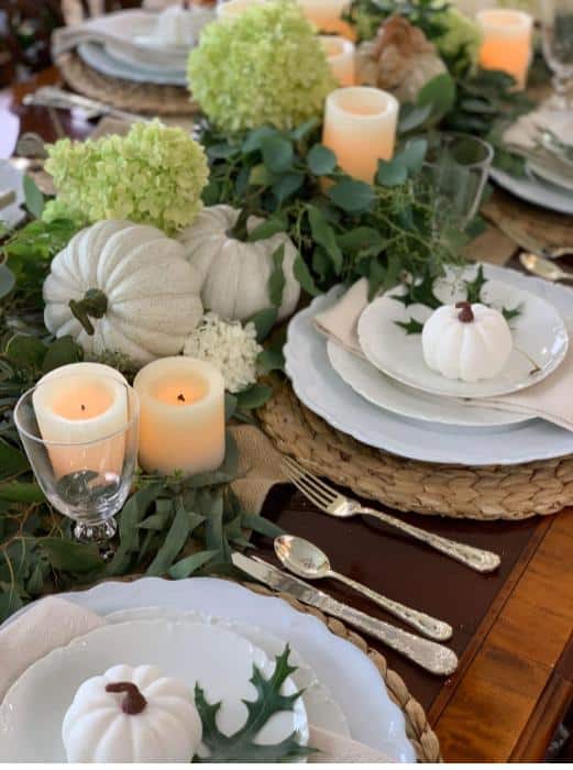 White fall table devor with natural woven placemants topped with white dinnerware, white napkin , green oak leaf and a white mini pumpkins. Down the center of the talbe is greenery topped with white pumpkins, candles and green hydrangeas.