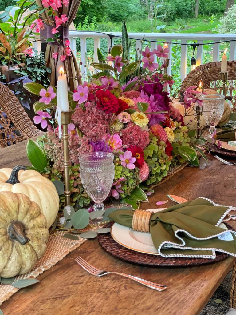 Outdoor fall table decor, rustic wood table with wicker chairs, burlap table runner with white and green pumpkins, brass candlessticks and a beautiful floral arrangment with cosmos, hydrangeas, zinnias, and assorted greenery. At each placesetting is a woven charger, and a gold rimmed white plate topped with an avocado green, lace trimmed napkin in a wooden napkin ring.