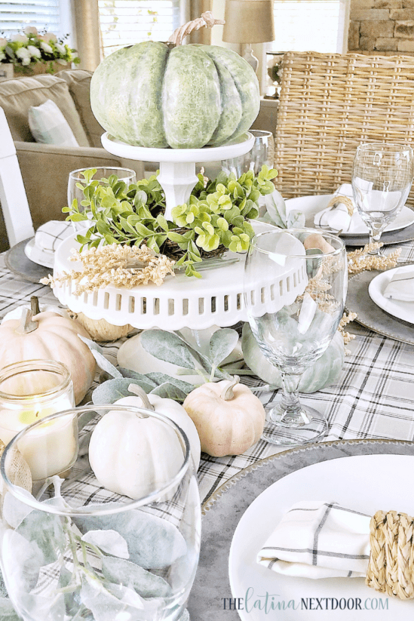 Farmhouse feel fall table decor, black and white plaid tablecloth, galvanized metal charger, topped with white places and a white and black plaid napkin in a woven napkin ring. Centerpiece is made with white milkglass pedestools topped with pumpkins and greenery, with more pumpkins scattered around the table