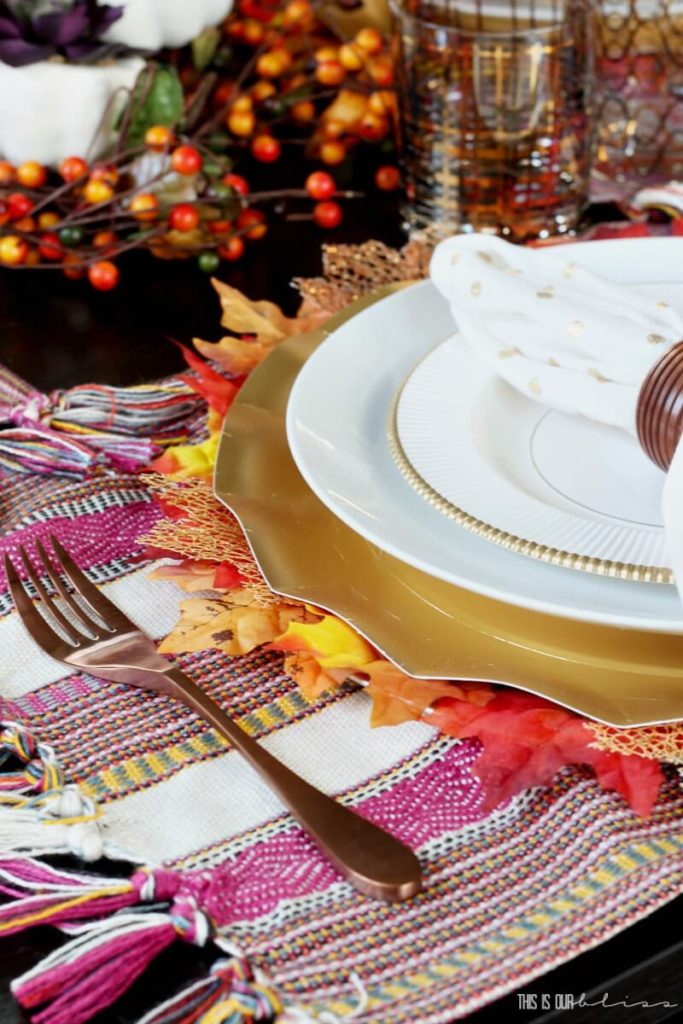 Bright and cheery fall table decor with a berry pink, yellow and white placemats topped with a leafs and a gold charger, White and gold rimmed plated topped with a white and gold napkin in a wooden napkin fing. Bittersweet berried are used for the centerpiece.