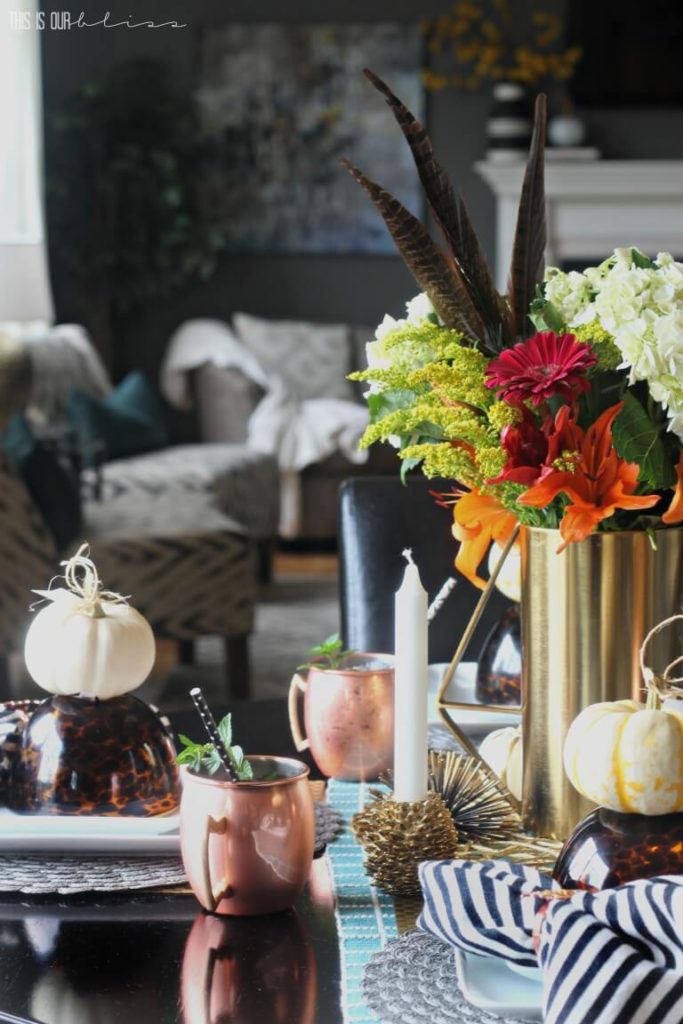 Fall tablescape with brass vase filled with white hydrangeas, orange lillies, goldenrod, feathers and red gerbera daisies. At each place setting is a square white place topped with a tortoise shell bowl and a white mini pumpkin. Copper mugs are placed at each place setting.