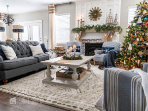 Living room decorated for Christmas, evergreen stems on the mantle with burlap stocks decorated with seashells, blue and white striped chairs, with blue and white snowflake pillos, white rug on hardwood floor, Christmas tree decorated with nautiful themed ornaments.