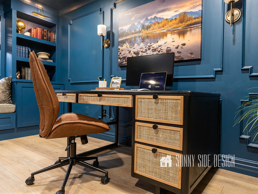 Home office library, with a mid-century modern desk with black stain, drawers have cane fronts with natural oak trim. On top of the desk dist a tray with accessories, a monitor and a laptop. Above the desk is another piece of photography artwork with 2 brushed brass wall sconces with white shades. Wall is painted blue with picture frame moulding.