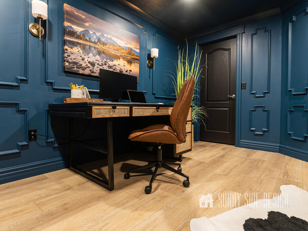 Cozy home office library with dark blue walls with picture frame moulding, landscape photography art, and brushed brass wall sconces. Against the wall is a mid-century modern black stained desk wiht cane and oak trim drawer fronts and a camel leather office chair.