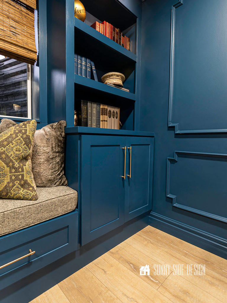 Built-in bookshelves, with brushed gold cabinet pulls, filled with books, blue wall with picture frame moulding, windowseat with brown herrinbone cushion with kilim pillows, window has a bamboo roman shade.