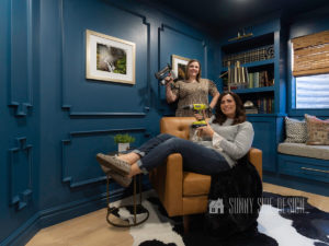 Home office library with 2 women holding power tools, one sits on a camel leather club chair and the other sands behind the chair, brass floor lamp, dark blue painted walls with picture frame moulding. Landscape photography art is framed and mounted to the wall, built-in bookshelves painted dark blue, filled with books and mementos, brass library light at the top of the bookshelves, Windowseat below window with brown cushion and kilim pillows.
