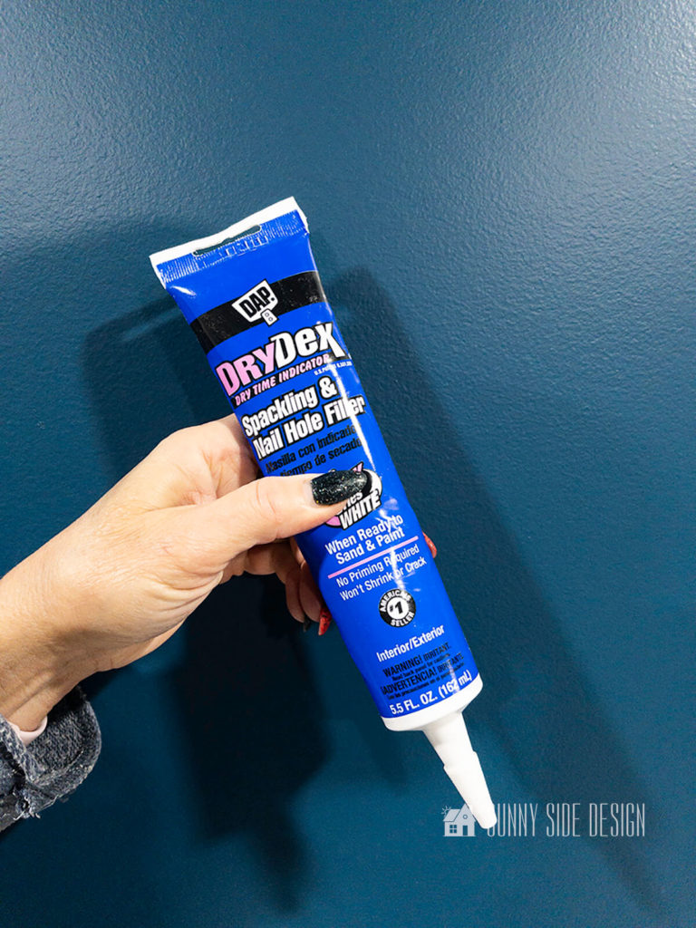 Caulk, Wood Filler or Spackle, woman holds a tuve of spackling nail hold filler against dark blue wall.