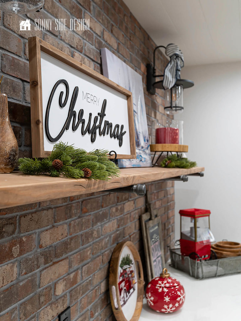 Effortless Holiday decorating ideas at the wet bar, frames "merry Christmas" sign on rustic wood shelf with greenery picks with pinecones, red candel on a bamboo stand and festive ribbon bows on the rustic lanterns on the rustic brick wall. On the epoxy counters is a red ornament cookie jar and a festive tray.