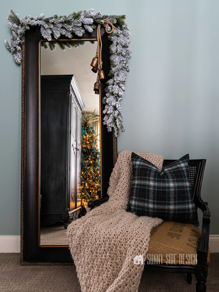 DIY Christmas bells hung on a black framed floor mirror with a flocked garland. In the reflection of the mirror your can see a lit Christmas tree and black armoire.