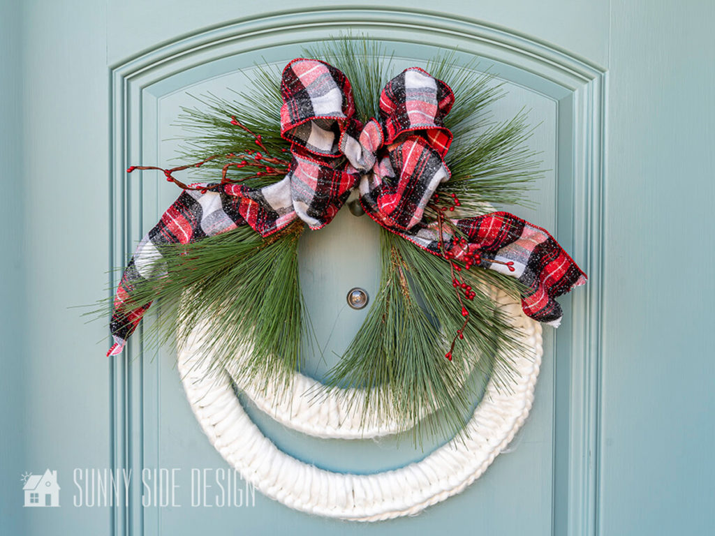 Chunky white yarn Christmas wreath embellished with greens and a flannel plaid bow on a light blue door.