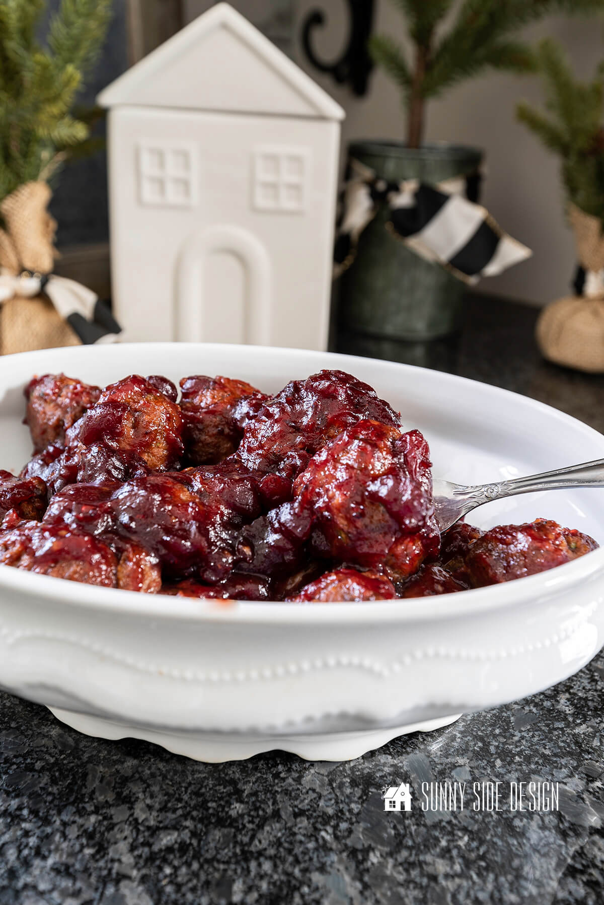 Easy meatball appetizer in a cranberry sauce in a white serving dish with fork. In the background are mini pine trees and a ceramic house.