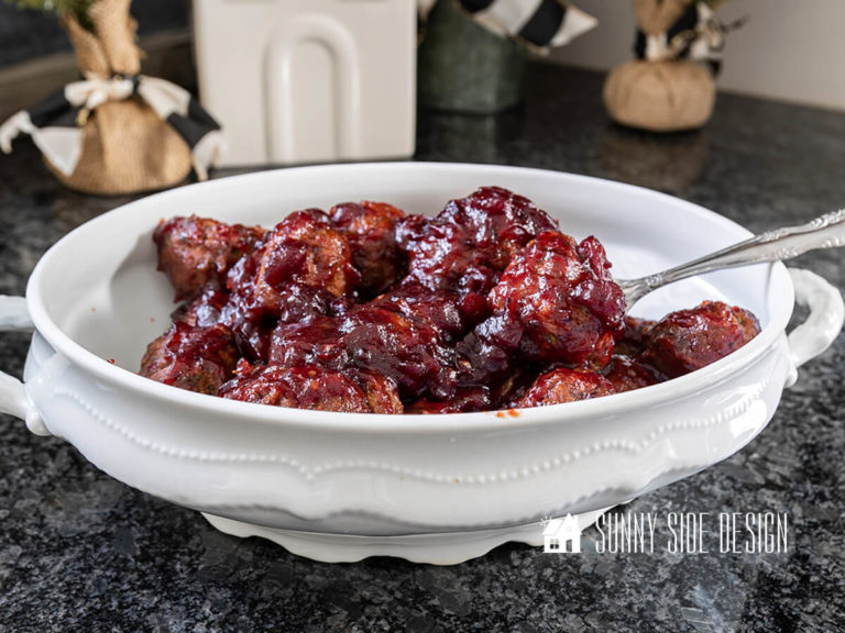 Easy meatball appetizer in a cranberry sauce in a white serving dish with fork. In the background are mini pine trees and a ceramic house.
