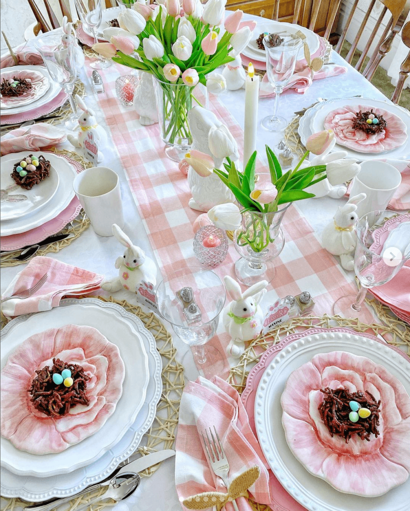 Pink and white themed Easter tablescape with a pink gingham table runner, pink and white layered dinnerware, pink and white tulips and small bunnies scattered around the table.