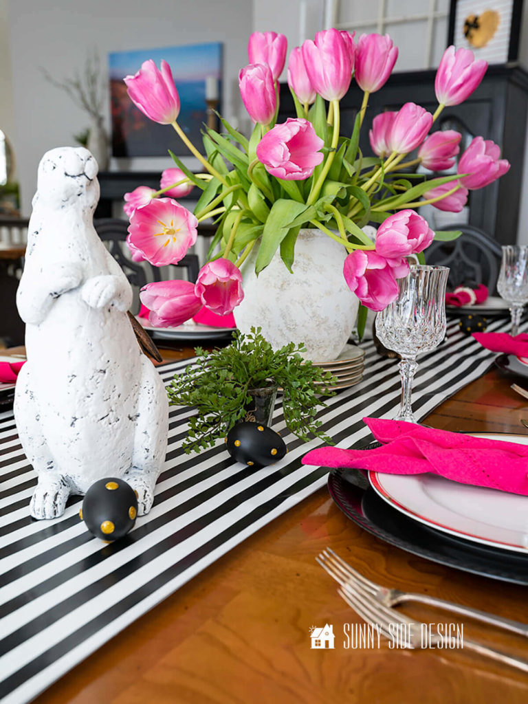 Kate Spade inspired Spring table decor ideas: black and white paper table runner, eggs embellished with gold dots, black chargers with gold rimmed dinnerware, bunnies, green plants and fresh hot pink tulips. Each place setting has a hot pink napkin folded into a bunny.