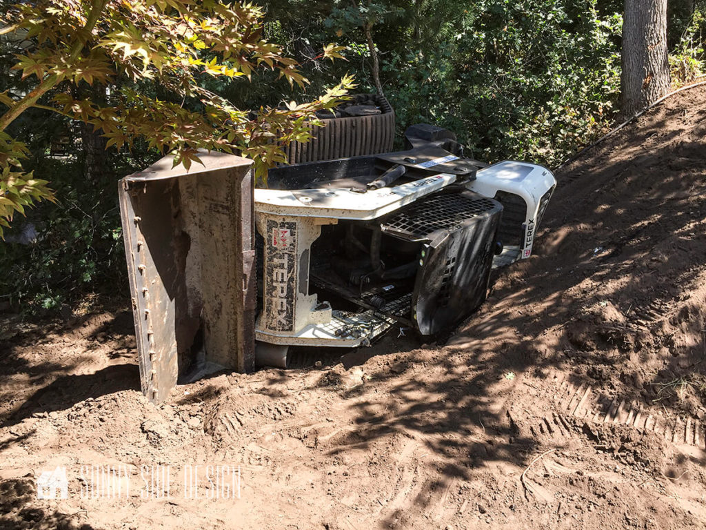 Skid loader tipped over on its side in wooded backyard.