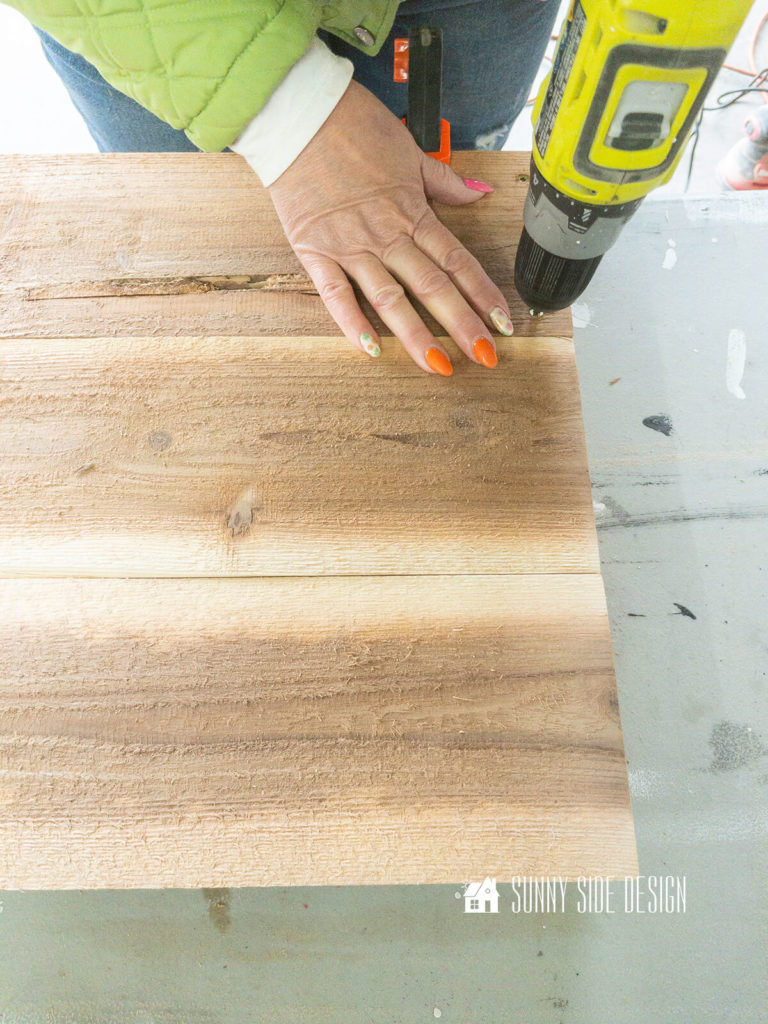 Woman holding a screw driver, secures cedar plank side boards with deck screws to the previous built cedar box sides.