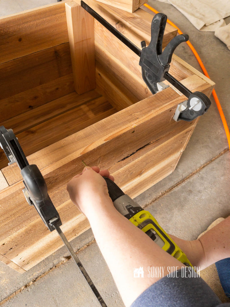 Two clamps are used to hold in place a piece of 2x4 cedar, while a woman places screw into the cedar box.