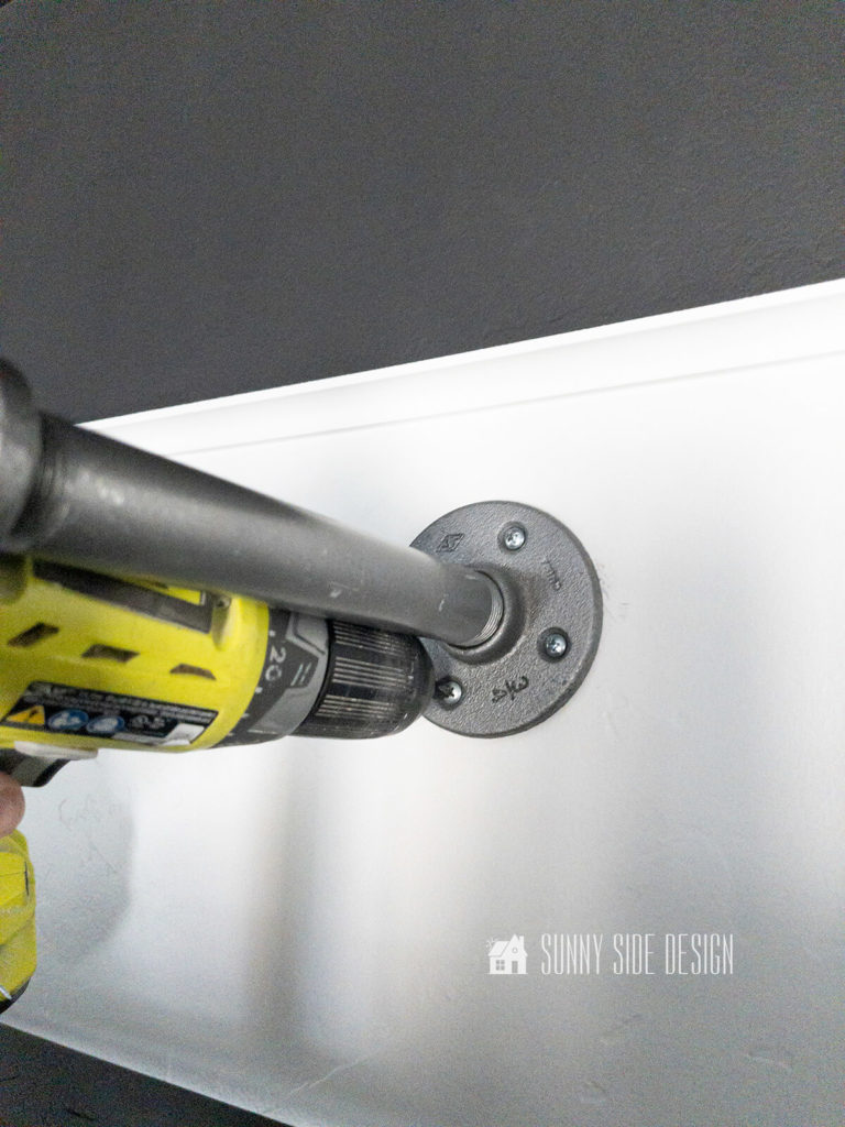 A drill is used to insert screws to secure the pipe flanges to the wall.