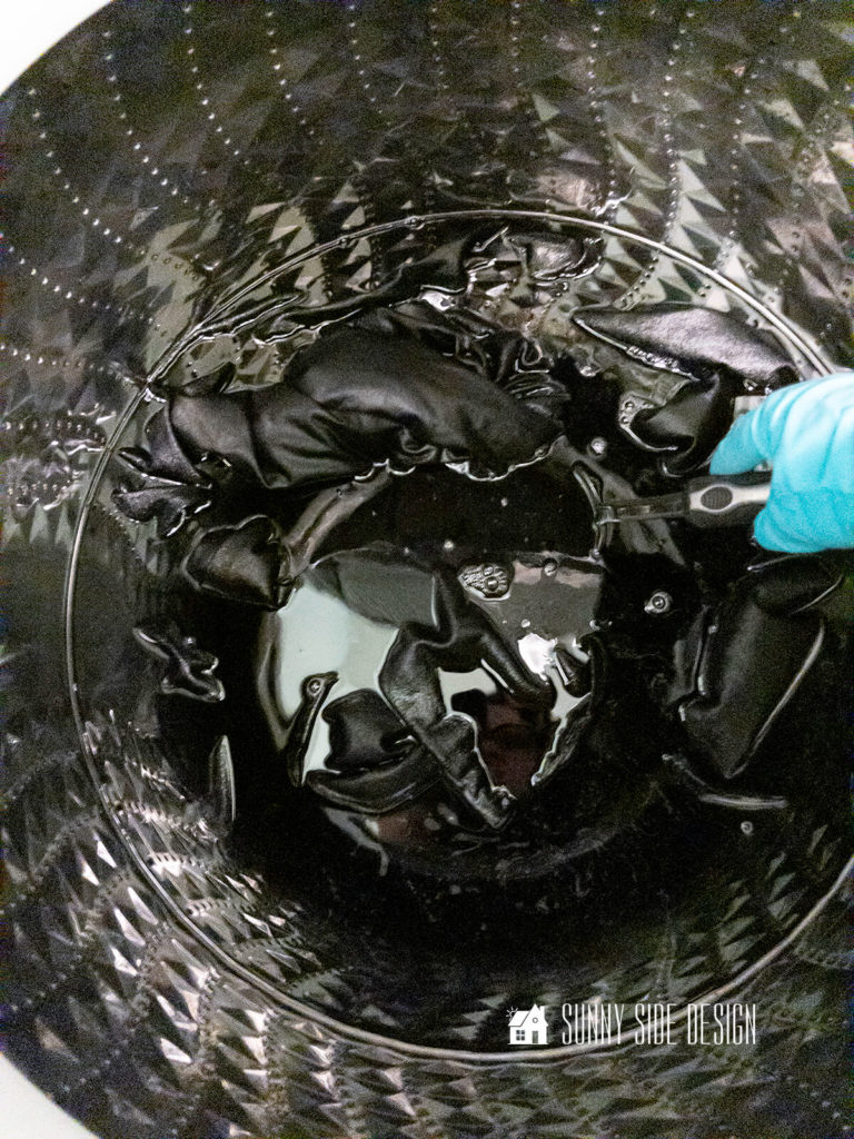 Black Rit Dye is added to washed fabric in a top loading washing machine and a woman wearing rubber gloves stirs the fabric with a stainless steel spoon.