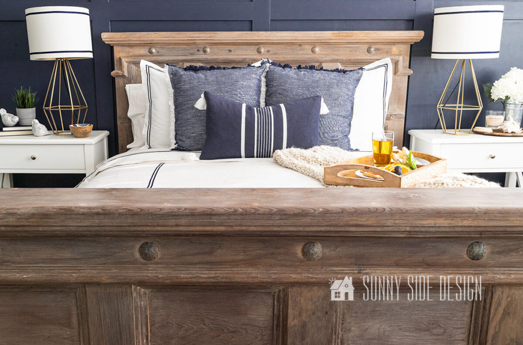 Newly refinished bed with an aged wood finish inspired by Restoration Hardware styled with white bedding embellished with navy blue trim. Bed against a navy blue board and batten wall with framed coastal photography. White nightstands with gold geometric lamps with white lampshades trimmed with navy blue grosgrain ribbon.