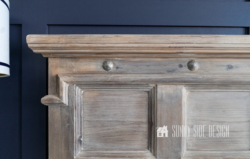 Close up imaged of refinished pine bed with an aged, weathered finish using vinegar and steel wool. Headboard is against a navy blue board and batten wall.