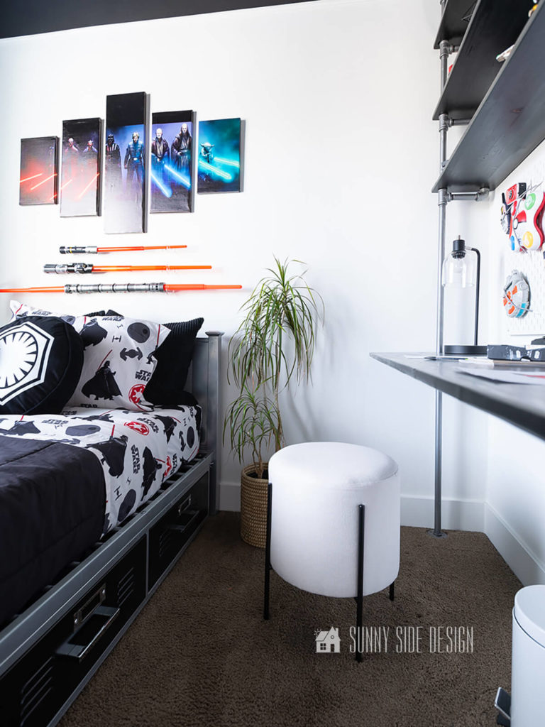 Star Wars small bedroom ideas, white walls with black ceiling, Star Wars canvas art is mounted above the bed along with 3 light sabers. Metal bed with black locker style drawers painted black and staged with a black comforter, Star Wars themed sheets and pillows. White stool with black legs is placed at the wood and pipe desk.