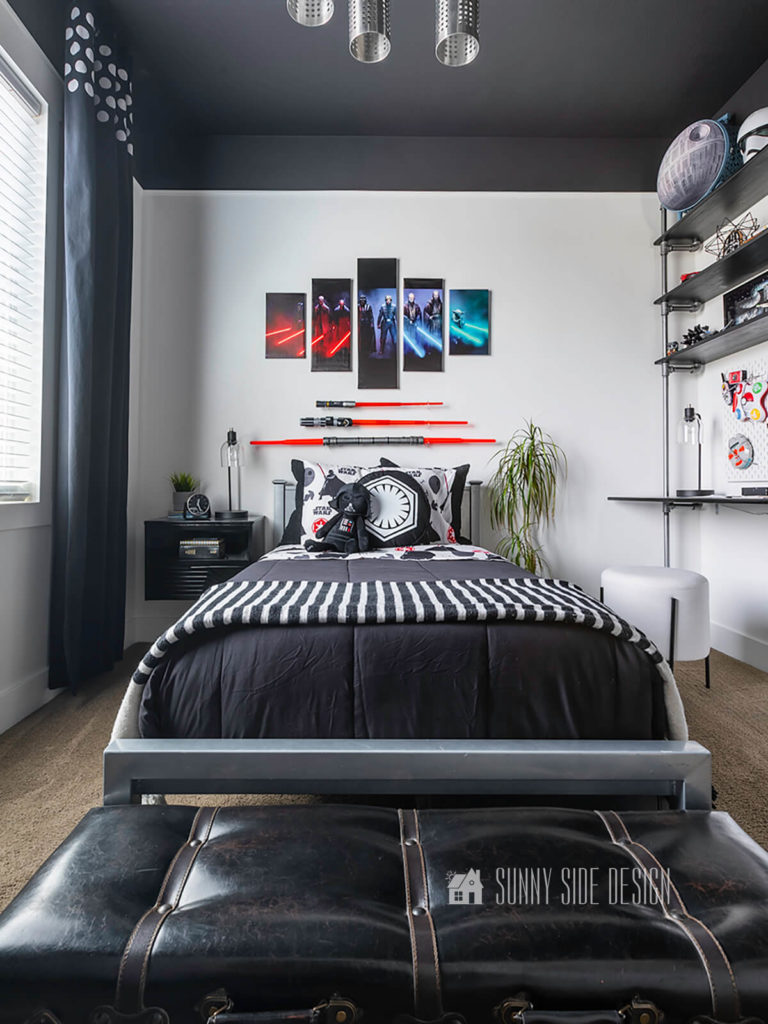 Star Wars small bedroom ideas, white walls with black ceiling, Star Wars canvas art is mounted above the bed along with 3 light sabers. Metal bed with black locker style drawers painted black and staged with a black comforter, Star Wars themed sheets and pillows. A black and white striped throw blanket is at the foot of the bed along with a Star Wars costume. White stool with black legs is placed at the wood and pipe desk. Black floating nightstand is to the right of the bed with a faux leather trunk at the foot of the bed.