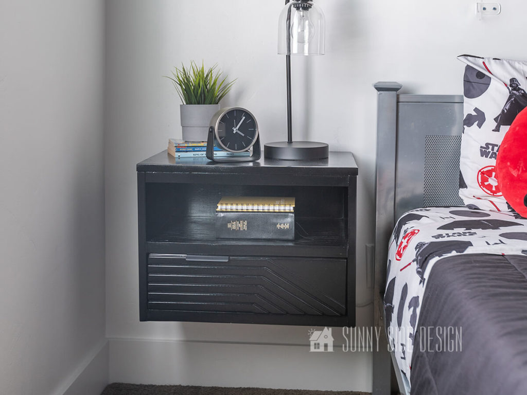 Modern black floating nightstand with design on drawer front with screen moulding. Black lamp, clock and potted plant on top of nightstand. Silver metal bed with black, white and red Star Wars Bedding.