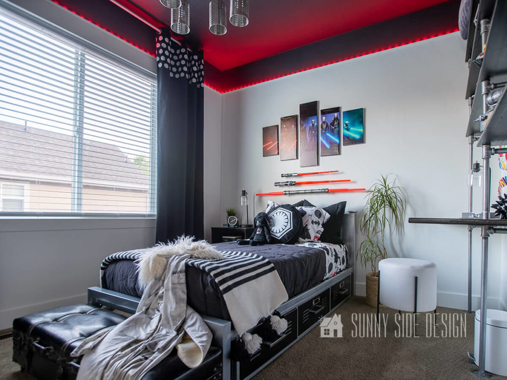 Star Wars small bedroom ideas, white walls with black ceiling, red LED lights encircle the room, Star Wars canvas art is mounted above the bed along with 3 light sabers. Metal bed with black locker style drawers painted black and staged with a black comforter, Star Wars themed sheets and pillows. A black and white striped throw blanket is at the foot of the bed along with a Star Wars costume. White stool with black legs is placed at the wood and pipe desk. Black curtains with white polka dot detailing at top.