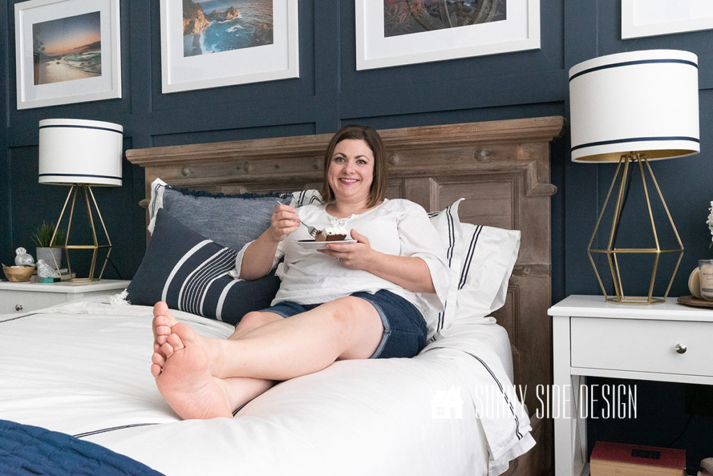 Woman sitting on newly refinished bed with an aged wood finish inspired by Restoration Hardware styled with white bedding embellished with navy blue trim. Bed against a navy blue board and batten wall with framed coastal photography. White nightstands with gold geometric lamps with white lampshades trimmed with navy blue grosgrain ribbon.