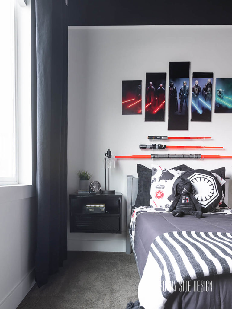 Star Wars Small Bedroom Ideas: black floating nightstand is mounted next to a metal bed with under drawer storage. Bed is decorated with black, white and red Star Wars bedding with a black comforter and a black and white throw blanket. Mounted above the bed are 3 red light sabers.