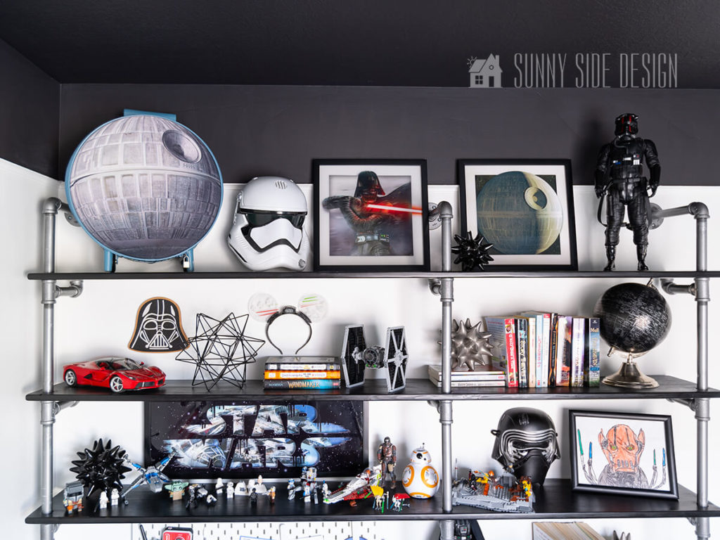 Wood and pipe display shelves with Star Wars themed art, trooper mask, star orbs, Star Wars death star, red RC car, novels, Lego Star Wars fighter, Darth Vader, black globe, Lego Star Wars action figures and child's Star Wars framed art.