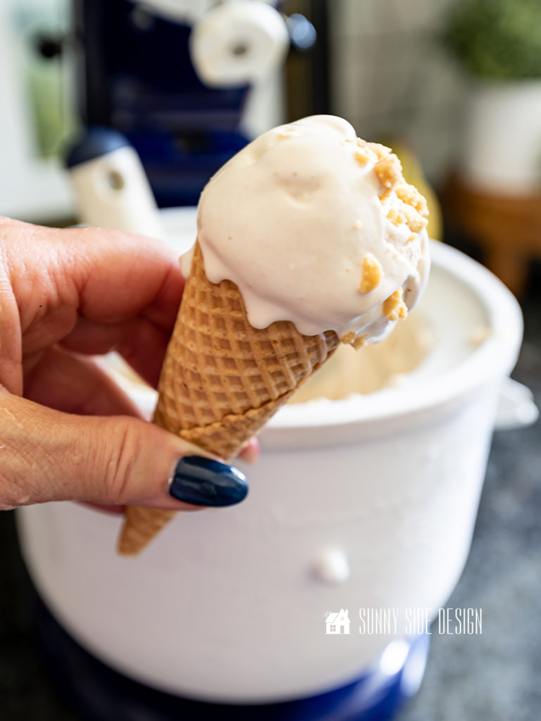 Peanut Butter Banana Homemade ice cream in a waffle cone sprinkled with chopped nuts in a woman's hand. In the background is a Kitchenaid ice cream maker attachment.