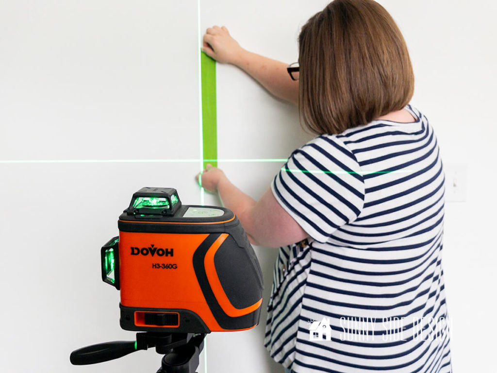 Laser level in the foreground with the laser beam projected on a white wall. Woman is placing painters tape along the laster beam.