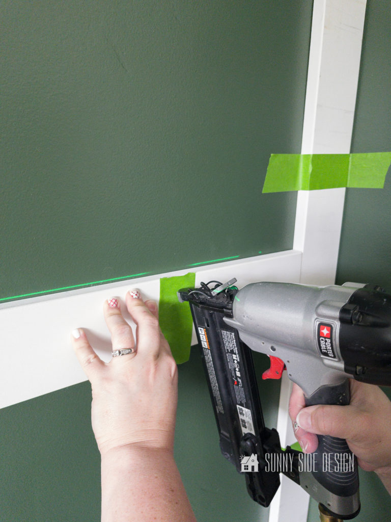 Woman's hand securing board to a green painted wall with a brad nailer.